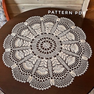 May include: Cream-coloured, circular doily with an intricate floral and geometric lace pattern. The doily is centered on a dark brown wooden table. The words "PATTERN PDF" are visible in the upper right corner.