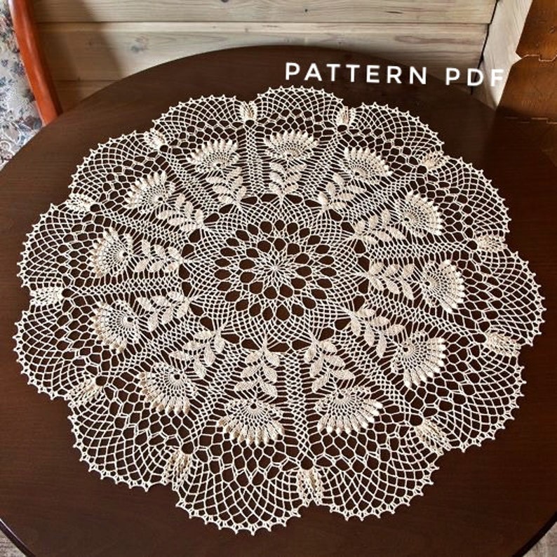 May include: Cream-coloured, circular doily with an intricate floral and geometric lace pattern. The doily is centered on a dark brown wooden table. The words "PATTERN PDF" are visible in the upper right corner.