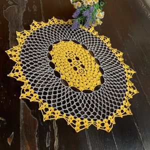 Op de afbeelding: Een geel en grijs gehaakt tafellaken met een cirkelvormig ontwerp. Het tafellaken heeft een delicaat, kantpatroon en is perfect om elke tafel een vleugje elegantie te geven.