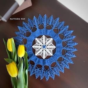 May include: A handmade blue and white crochet doily with a starburst design. The doily is circular, with a detailed openwork pattern. The text "PATTERN PDF" is visible in the upper left corner. A bouquet of yellow tulips is in the lower left corner.