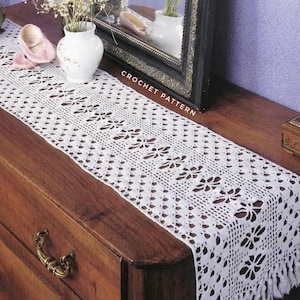 May include: A white, crocheted table runner with a floral pattern and fringe detail. The runner is displayed on a dark wooden dresser. The words "CROCHET PATTERN" are visible in the image.
