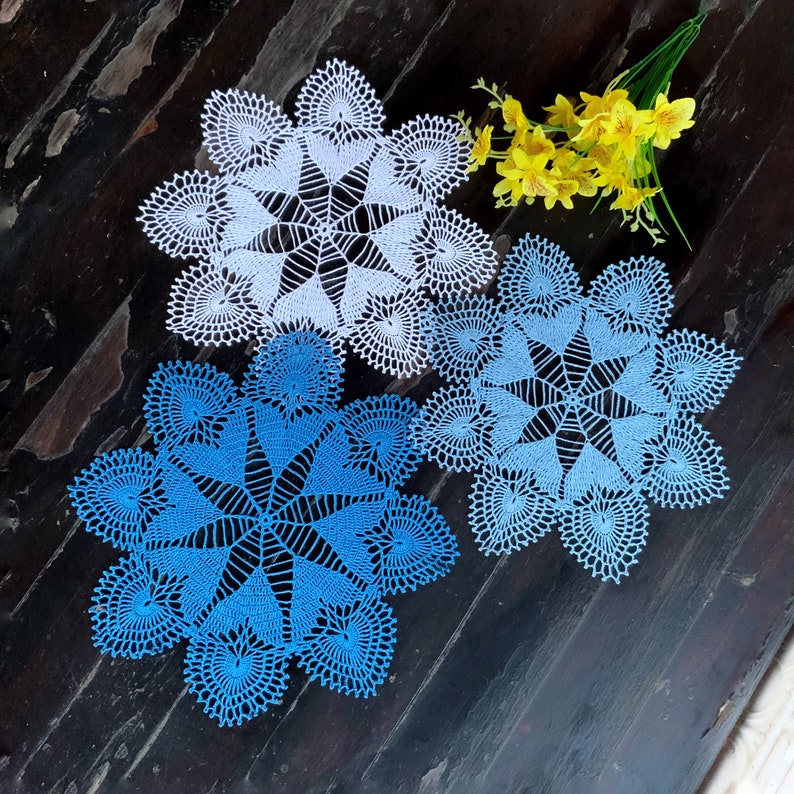 May include: Three crocheted doilies, one white and two blue, with intricate patterns. The doilies are arranged on a dark wooden surface.