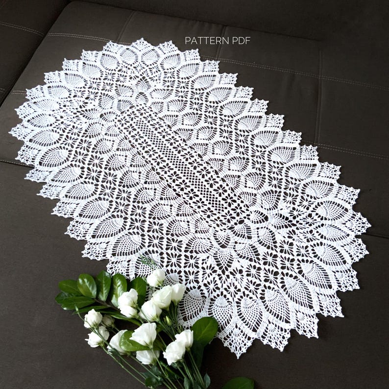 Pu&ograve; includere: Un tovaglia ovale in pizzo bianco con un motivo delicato e intricato. La tovaglia &egrave; stesa su una superficie marrone e presenta un motivo ripetitivo di piccoli fiori e foglie.