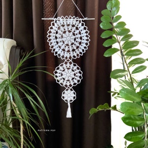 May include: White crocheted wall hanging with a geometric pattern. The hanging is made up of three circular pieces, each with a different design. The hanging is attached to a wooden dowel rod and has a tassel at the bottom.