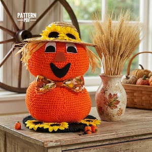 May include: A crochet scarecrow figure with a bright orange body, a straw hat adorned with yellow sunflowers, and a patterned bow tie. The figure features black eyes, a triangular nose, and a wide smile. Text on the image reads PATTERN PDF.