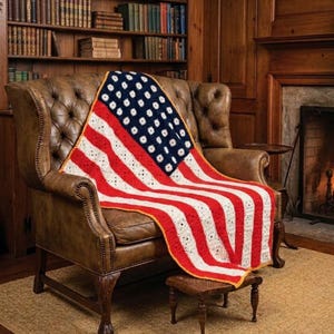Pode incluir: Uma manta de crochê com o design da bandeira americana, drapeada sobre uma poltrona de couro marrom. A manta tem listras vermelhas e brancas e uma seção azul com estrelas brancas. Possui uma borda amarela.
