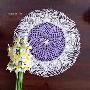 May include: A handmade, white and lavender crochet doily with a floral design. The doily is round and has a detailed, openwork pattern. The text "PATTERN PDF" is in the upper left corner. A bouquet of white and yellow daffodils is in the lower left corner.