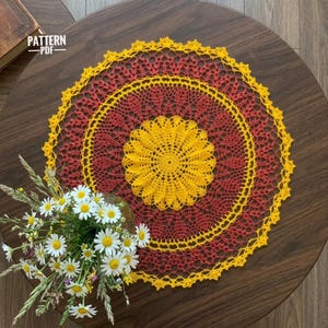 May include: A round, intricate crochet doily featuring a vibrant yellow center and outer border, with a deep red circular pattern in between. The doily has a scalloped edge and is displayed flat, accompanied by a bouquet of white daisies and green grass.