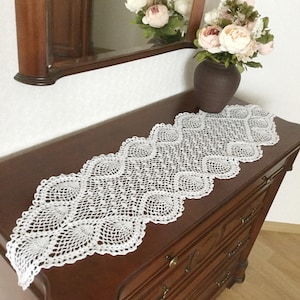 Peut inclure: Chemin de table blanc, finement crocheté, avec un bord festonné, présenté sur une commode en bois foncé. Le motif ajouré du chemin de table est visible. Un vase de fleurs et un miroir sont en arrière-plan.
