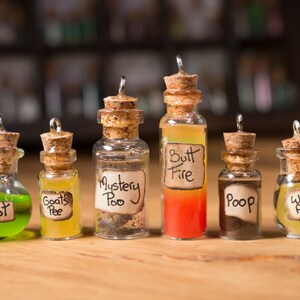 May include: Seven small glass bottles with cork stoppers and labels. The bottles contain different colored liquids and have humorous labels such as "Snot", "Goat's Pee", "Mystery Poo", "Butt Fire", "Poop", and "Wolf Pee".