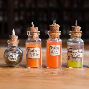 May include: Six small glass bottles with cork stoppers and metal caps, each filled with a different coloured liquid and labelled with handwritten text. The labels read "Virus", "Wolfs Bane", "Radio-Active", "Mystical Virus", "Radioactive Virus", and "Fart Gas".
