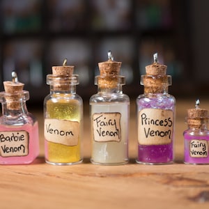 Puede incluir: Cinco pequeñas botellas de vidrio con tapones de corcho y tapas plateadas. Cada botella contiene un líquido de color diferente y tiene una etiqueta escrita a mano con el texto "Venom" y una palabra diferente delante. Las etiquetas son: "Barbie", "Venom", "Fairy", "Princess" y "Fairy".