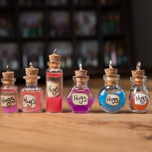 May include: A set of small glass bottles with corks, each containing a different coloured liquid. The bottles have labels saying "Hugs". The liquids are various colours, including pink, red, purple, blue, and orange. The bottles are on a wooden surface.