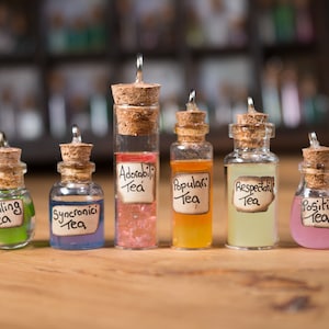 May include: Seven small glass bottles with cork stoppers and metal caps, each filled with a different colored liquid. The bottles have labels with handwritten text that reads "Healing Tea", "Synchronici Tea", "Adoptabili Tea", "Populari Tea", "Respectable Tea", and "Positivi Tea".