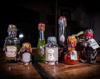 May include: A collection of six glass bottles filled with colored liquids, each sealed with dripping wax in various colors. The bottles feature vintage-style labels with text including 100% Health, Luck Potion, Extra Life, Unicorn's Milk, Love Potion, Dragon's Breath, and Beauty Poison.