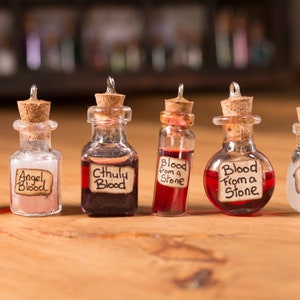 Könnte beinhalten: Sieben kleine Glasflaschen mit Korkstopfen und Etiketten. Die Etiketten sind handgeschrieben und sagen "Werwolfblut", "Engelblut", "Cthuly Blut", "Blut aus einem Stein", "Blut aus einem Stein" und "Einhornblut".
