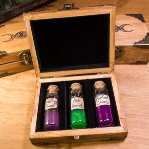 Puede incluir: Una caja de madera con tapa abatible, que contiene tres pequeñas botellas de vidrio con tapones de corcho. Las botellas están llenas de líquidos morado, verde y morado. Las etiquetas de las botellas dicen "Fieling Power", "Anti Anxiety" y "Bottled Chaos".