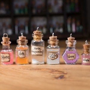 May include: Seven small glass bottles with cork stoppers and silver caps. Each bottle contains a different coloured liquid and has a handwritten label that says "Hope" in various styles. The bottles are arranged in a row on a wooden surface.