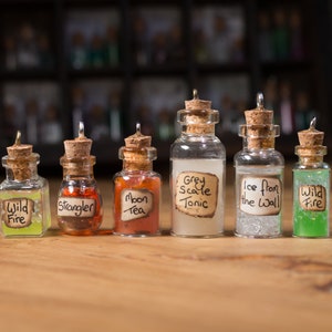 May include: Six small glass bottles with cork stoppers and handwritten labels. The labels read "Wild Fire", "Strangler", "Moon Tea", "Grey Scale Tonic", "Ice from the Wall", and "Wild Fire".