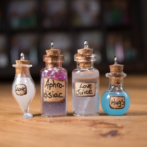 May include: Four small glass bottles with cork stoppers and handwritten labels. The labels read "Love", "Aphrodisiac", "Love Juice", and "Hugs". The bottles are filled with different coloured liquids.
