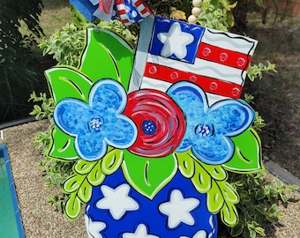May include: A blue and white vase with white stars painted on it. The vase is filled with red, white, and blue flowers. A red, white, and blue bow is attached to the top of the vase. The vase is sitting on a bed of gravel.