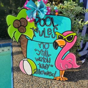 May include: A turquoise wooden sign with the words "Pool Rules" and "No "y'all watch this" allowed" in white paint. A pink flamingo with black sunglasses and a beach ball are on the sign.