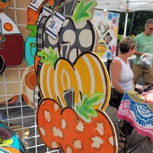 May include: A three-tiered pumpkin door hanger with a black and white polka dot pumpkin on top, a yellow and white striped pumpkin in the middle, and an orange pumpkin with a leopard print on the bottom. Each pumpkin has a green stem and leaves.