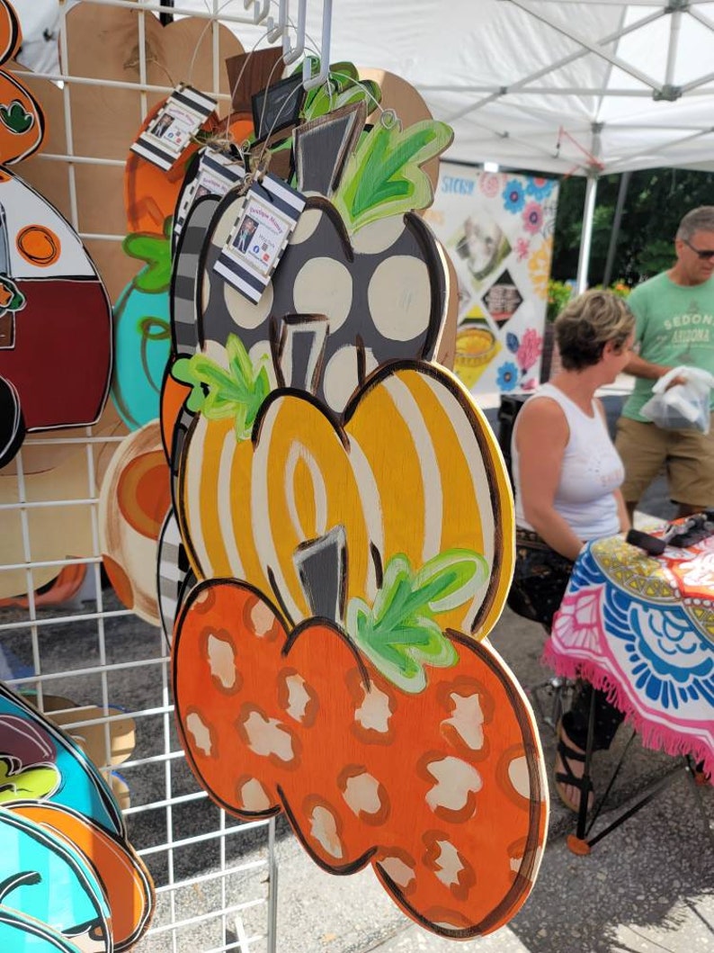 Puede incluir: Un colgador de puerta de calabaza de tres niveles con una calabaza de lunares negros y blancos en la parte superior, una calabaza rayada amarilla y blanca en el medio y una calabaza naranja con estampado de leopardo en la parte inferior. Cada calabaza tiene un tallo y hojas verdes.