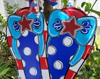 May include: A pair of blue and red flip flops with white polka dots and red stars. The flip flops are decorated with a blue and white bow and a red, white, and blue bow. The flip flops are hanging on a tree branch.