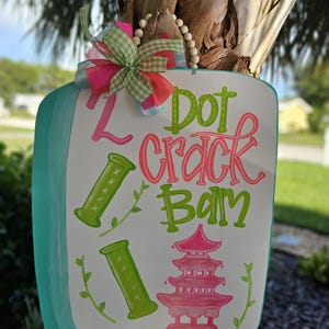 May include: A rectangular wooden sign with a turquoise border and the words "Z Dot Crack Bam" in green and pink. The sign features green bamboo-like shapes and a pink pagoda design. A decorative bow with pink, green, and white ribbons adorns the top.