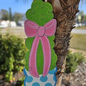 May include: A green bush with a pink bow and blue polka dot pot. The bush is hanging from a wooden bead garland.