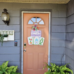 May include: A peach-colored front door with a decorative sign that reads "Mahjong is My Favorite Spot." The sign features three Mahjong tiles. A green and purple bow is above the sign. The door is surrounded by gray siding and green plants.