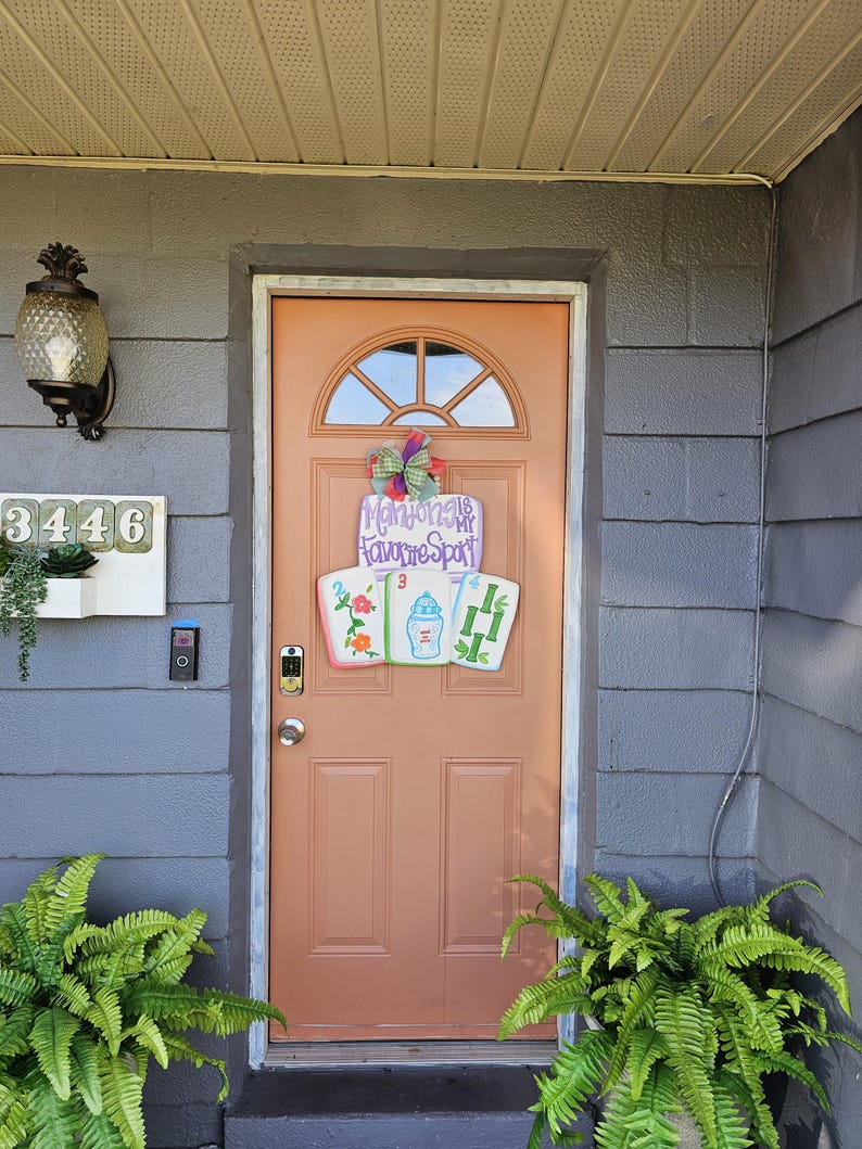 May include: A peach-colored front door with a decorative sign that reads "Mahjong is My Favorite Spot." The sign features three Mahjong tiles. A green and purple bow is above the sign. The door is surrounded by gray siding and green plants.