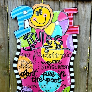 May include: A pink and white sign with the word "POOL RULES" in colorful letters. The sign has a sun, a beach ball, and a striped design. The text on the sign reads: "MAKE friends & MEMORIES, FLIP FLOPS are OPTIONAL, wear SUNSCREEN, don't pee in the pool, Enjoy the FUNNIN', NO RUNNIN', LOTSA SUNNIN'."