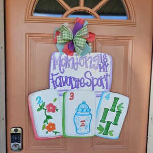 May include: A wooden door sign with the text "Mahjong is my Favorite Sport" in purple script. The sign features three painted Mahjong tiles and a colorful bow. The tiles are white with painted designs and numbers.