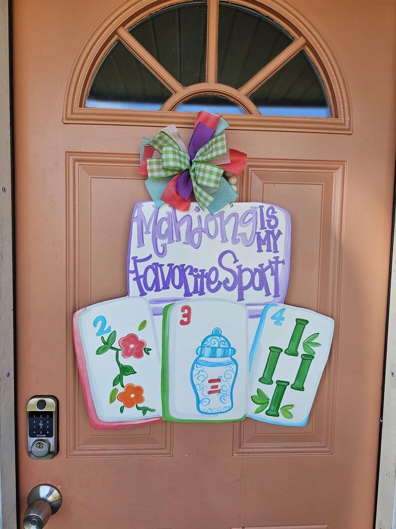 May include: A wooden door sign with the text "Mahjong is my Favorite Sport" in purple script. The sign features three painted Mahjong tiles and a colorful bow. The tiles are white with painted designs and numbers.