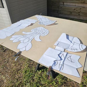 May include: A light brown wooden surface with several white paper cutouts of various shapes, including flowers and abstract designs. The cutouts are held in place with metal clamps. The letters "PH" and other markings are visible on one cutout.