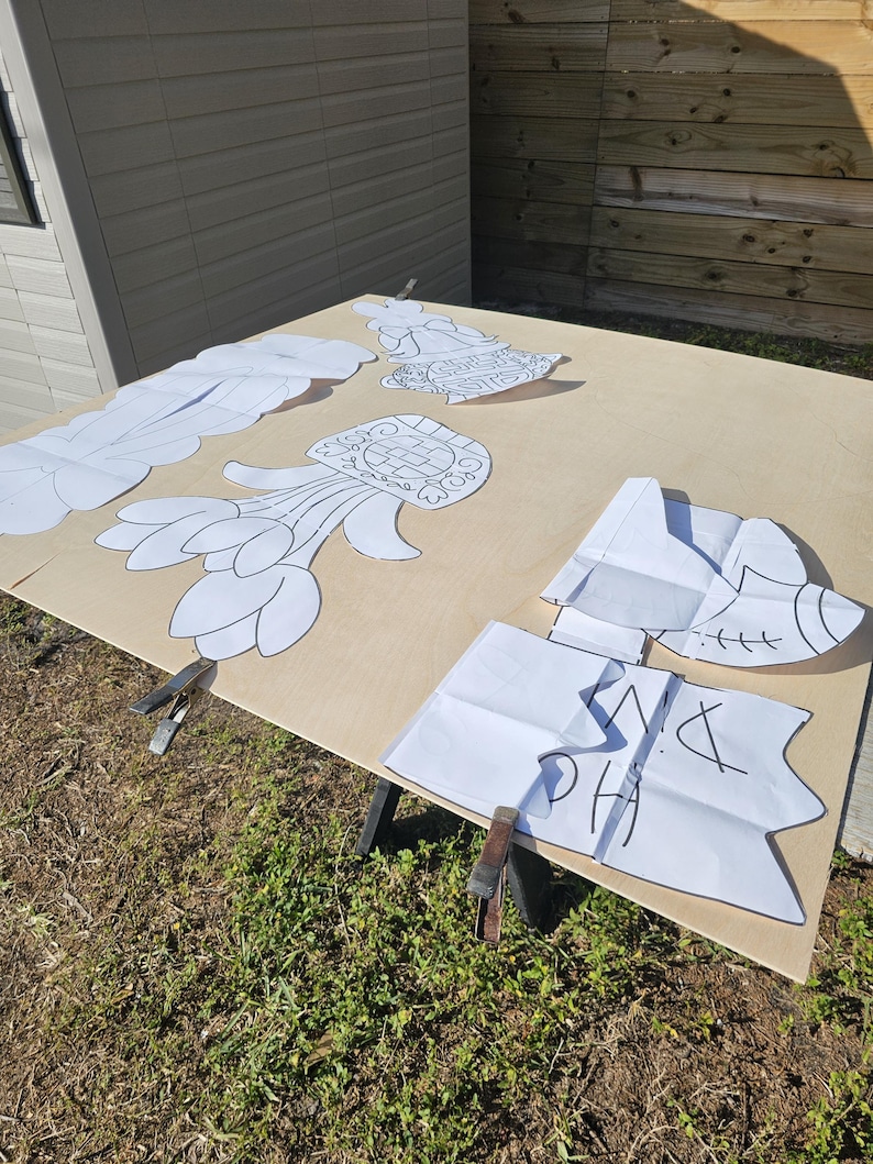 May include: A light brown wooden surface with several white paper cutouts of various shapes, including flowers and abstract designs. The cutouts are held in place with metal clamps. The letters "PH" and other markings are visible on one cutout.
