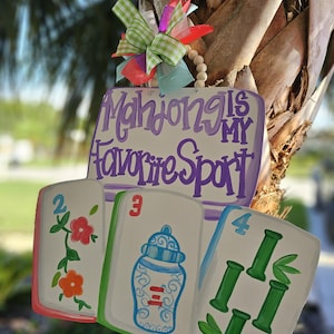 May include: A wooden sign with the text "Mahjong is My Favorite Sport" in purple script. The sign features three oversized mahjong tiles with colorful illustrations and a decorative bow.