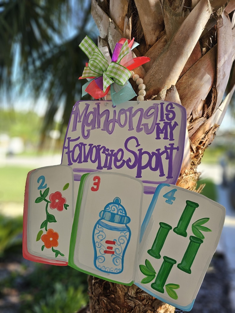 May include: A wooden sign with the text "Mahjong is My Favorite Sport" in purple script. The sign features three oversized mahjong tiles with colorful illustrations and a decorative bow.
