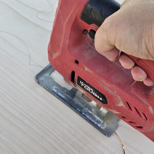 May include: A red and black Hyper Tough jigsaw cutting a light-colored wooden surface. The saw is in use, with the blade positioned to cut along a curved line. The image shows a close-up of the tool and the wood.
