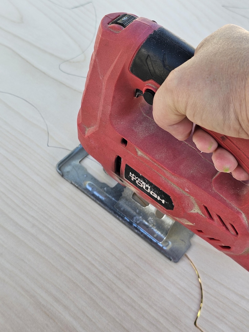 May include: A red and black Hyper Tough jigsaw cutting a light-colored wooden surface. The saw is in use, with the blade positioned to cut along a curved line. The image shows a close-up of the tool and the wood.