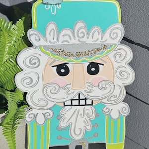 May include: A wooden Christmas nutcracker decoration. The nutcracker has a turquoise hat and uniform with white accents, a white beard, and a pink blush. A decorative bow with glitter and polka dots is attached to the top.