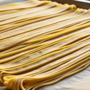 Pode incluir: Close-up de massa fresca e crua, disposta em fileiras paralelas numa assadeira. A massa é de cor amarelo claro, indicando que é fresca. A imagem destaca a textura e a forma da massa, mostrando fettuccine caseiro.