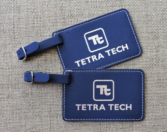 Puede incluir: Dos etiquetas de equipaje de cuero azul con costuras blancas. Las etiquetas tienen un logotipo blanco con las letras "Tt" dentro de un cuadrado y el texto "TETRA TECH" debajo del logotipo.