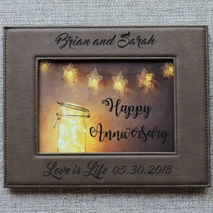 May include: A brown leather picture frame with a photo of a mason jar with lights inside. The frame is engraved with the text "Brian and Sarah", "Happy Anniversary", and "Love is Life 05.30.2018".