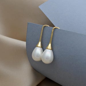 Op de afbeelding: Een paar goudkleurige oorhangers met peervormige, witte parelhangers. De oorbellen hebben een eenvoudig, elegant ontwerp, met een gebogen haak en een druppelvormige parel. De achtergrond is een mix van grijs en beige.