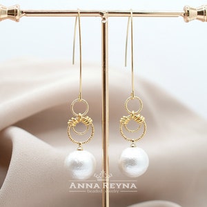 Op de afbeelding: Gouden oorhangers met een witte parel en twee gouden ringen. De ringen zijn met elkaar verweven en hebben een getextureerd ontwerp.