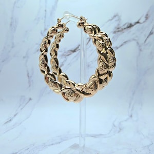 May include: Gold-colored hoop earrings with a braided design. The earrings are displayed on a clear stand against a marble-patterned background. The hoops have a textured surface, adding visual interest to the jewelry.
