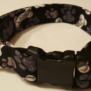 May include: Black dog collar with a white paw print and bone pattern. The collar has a black plastic buckle.
